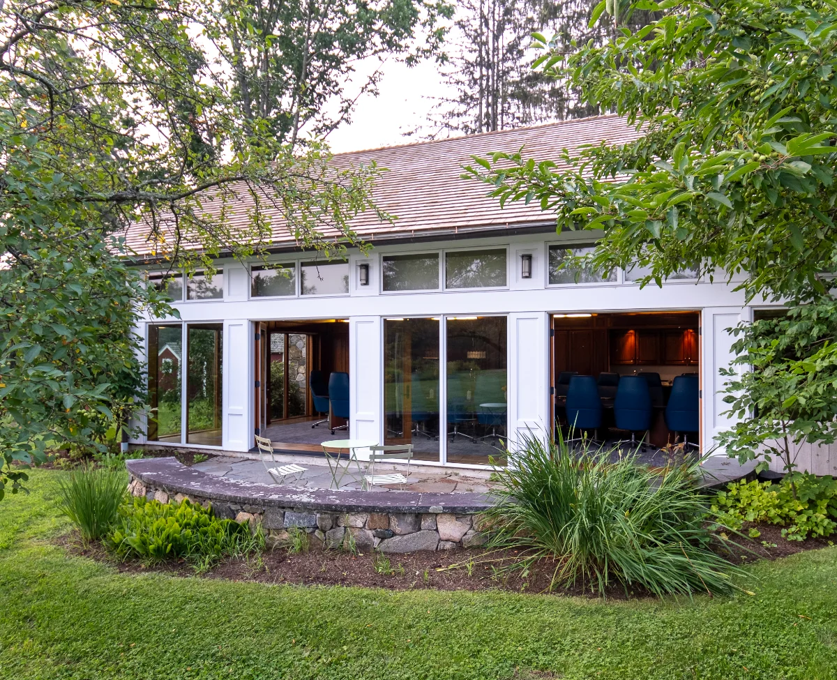 Exterior view of a modern cottage with large glass doors and windows, surrounded by greenery and trees