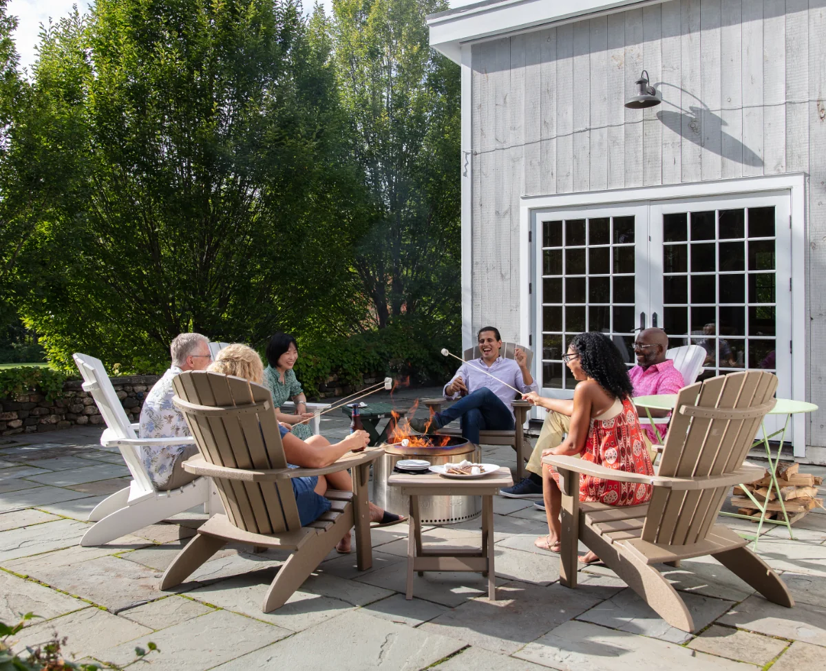 A group of people sitting in Adirondack chairs around a fire pit, roasting marshmallows.